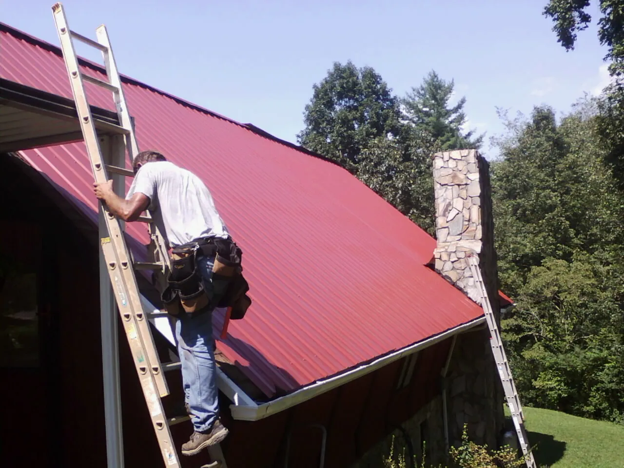 Roofer on a ladder beside a red metal roof and stone chimney — Soffit & Fascia Repair by Roofing Brothers in Little Elm